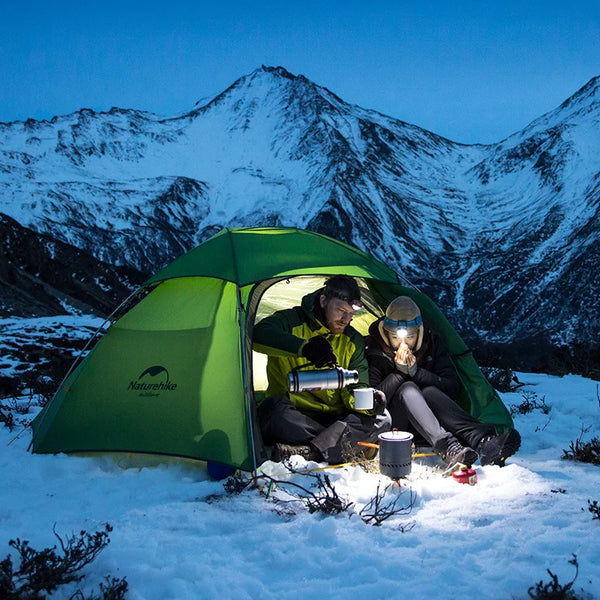 Un homme et une femme sont dans une tente dans la nature sous la neige. La tente est verte. L'homme sert un café. 