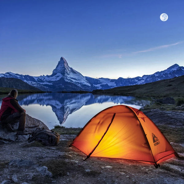Une tente orange igloo est installée devant un lac de montagne. Il y a une femme assise à côté.