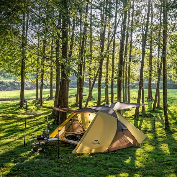 C'est une tente allongée deux places  moutarde et marron. Elle est posée dans un espace vert entourée  d'arbres. 