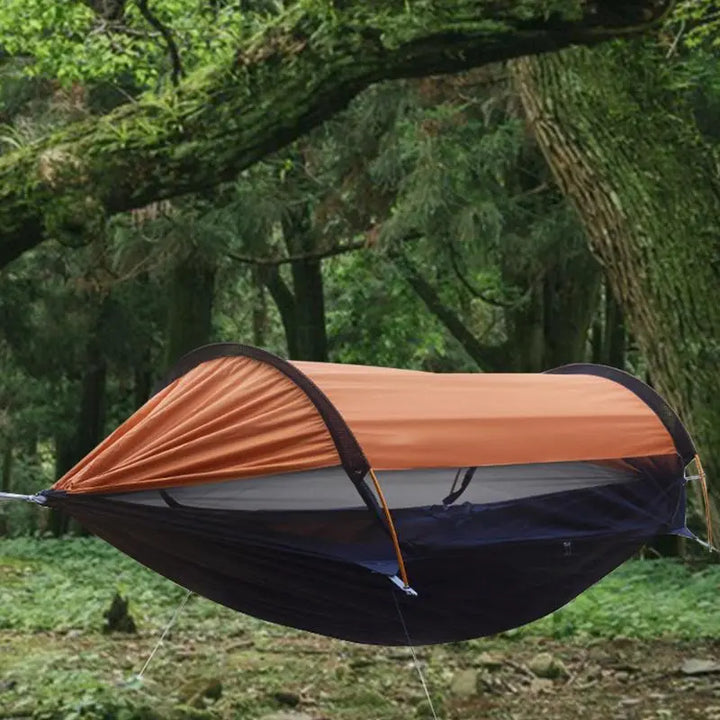 Une tente suspendue noire et orange est accrochée à des arbres dans une forêt. La tente est anti uv et avec une moustiquaire. 