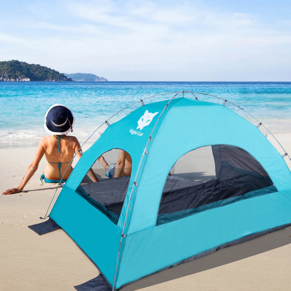 Une tente de plage anti uv bleu est posée sur une plage. Il y a un couple devant qui est assis dans le sable. 