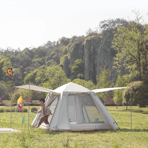 Une tente avec 4 grandes portes est installée sur une étendue d'herbe. La tente est beige. Un enfant sort de la tente. 