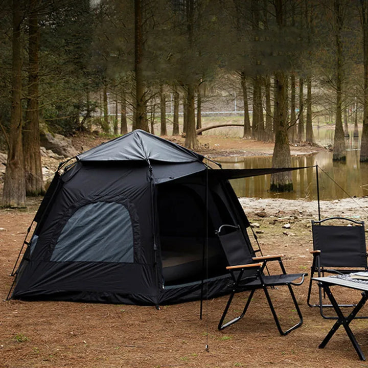 Une tente de camping noir est installée dans une forêt. Elle a un auvent et des fenêtres. Devant la tente il y a une table et des chaises. 