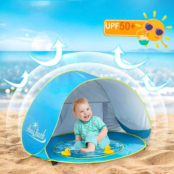 Un bébé est installée dans une tente de plage anti uv avec piscine sur le sable avec la mer derrière. La tente est bleue.