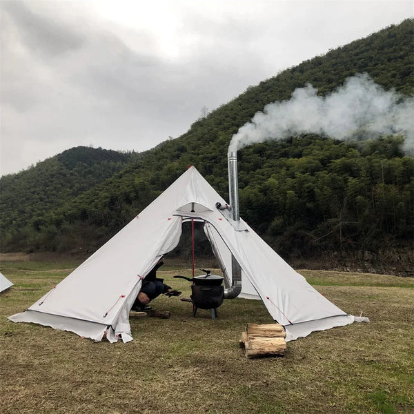 Une tente tipi blanche est posée dans un champ. Elle est équipée d'une cheminée avec un poêle à l'intérieur. Il y a du bois posés devant la tente. 