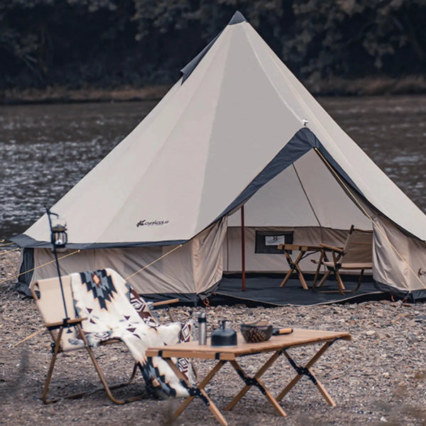 Une tente berbère beige et bleue familiale est installée au bord d'une rivière. Il y a une table basse et un fauteuil est devant la tente. 