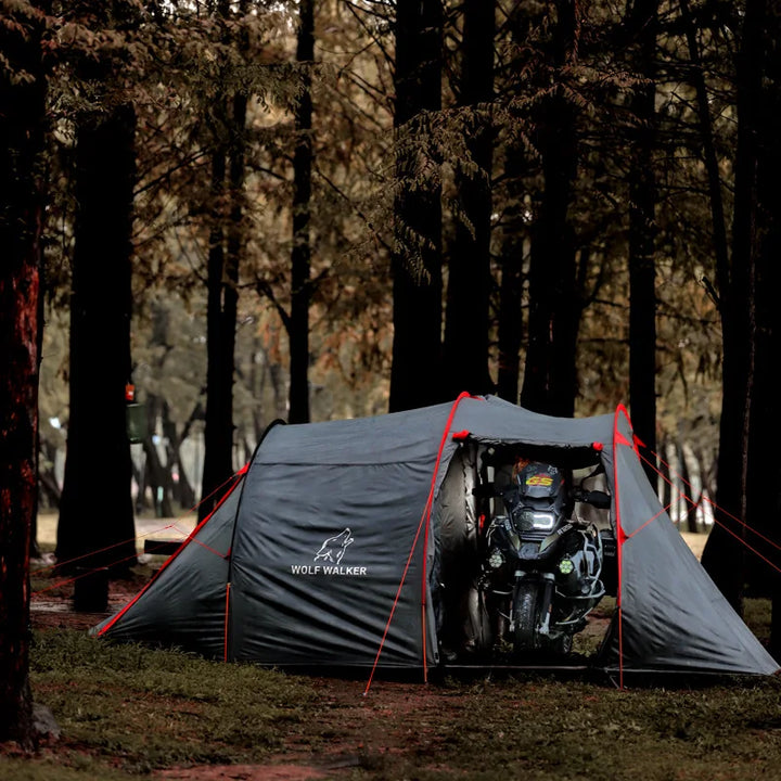 Une tente tunnel grise est installée en forêt. C'est une tente avec une chambre et un abri pour la moto. 