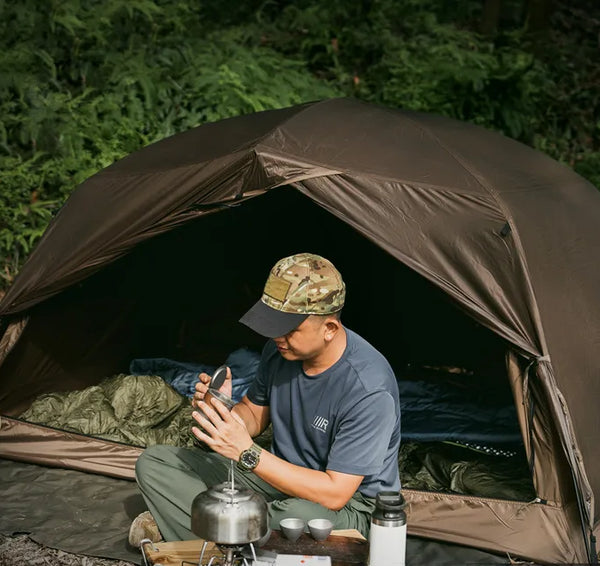 Un homme est assis devant une tente marron igloo. La tente est dans la nature. 