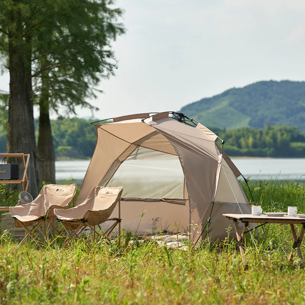 Une tente semi-ouverte de plage couleur beige est dans l'herbe à côté d'un étang. Il y a autour une table basse et deux fauteuils.