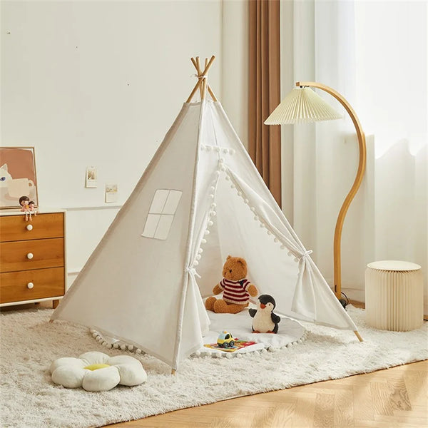 Une tente tipi indienne blanc avec pompons est installée dans une chambre d'enfant. La chambre est décorée en blanc et bois. Il y a des peluches dans la tente.