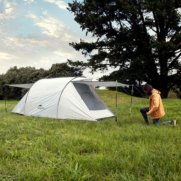 Une tente blanche avec deux auvents est installée dans une champ. Il y a un homme qui installe la tente et des arbres en fond. 