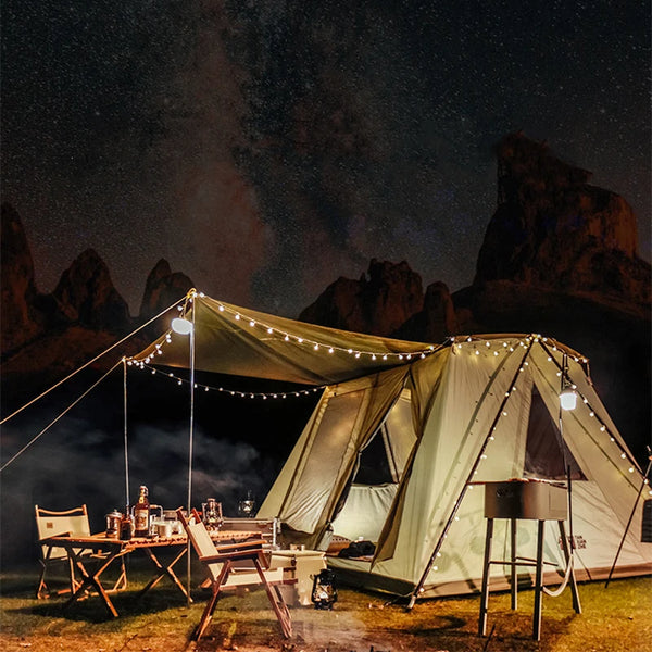 Une tente safari verte est installée dans la nature. Il fait nuit, elle est éclairée grâce à une guirlande lumineuse. Il y a sous le auvent de la tente une table, des chaises. Il y a également un barbecue à côté de la tente. 