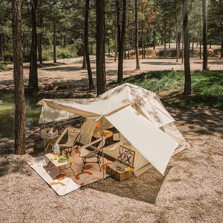 C'est une tente de camping avec un abri devant et une double porte. Elle est beige et est installée dans la nature avec des chaises et une table devant. 