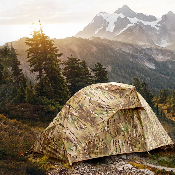 Une tente de trekking ultra légère militaire 1 place est installée dans une champ avec vue sur les montagne. La tente est dans un imprimé camouflage. 
