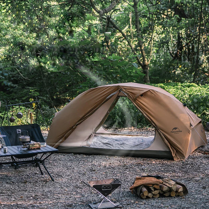 Une tente est installée dans la nature. Elle est marron avec une double porte. Il y a autour une table avec des chaises et un petit feu devant.  