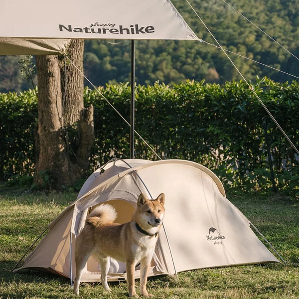 C'est une tente allongée beige pour chien. Elle est posée dans un jardin avec un chien devant. 