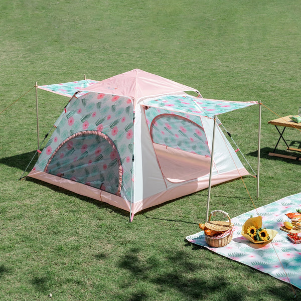 Une tente pour enfant rose avec un motif fleuri est installée dans un espace vert. La tente a deux portes et un auvent devant chaque porte. 