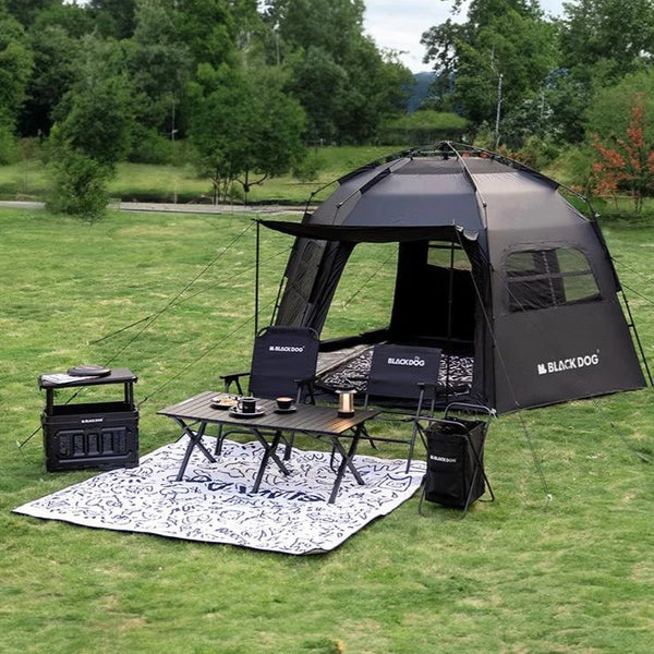 Une tente de camping familiale noire est installée dans un espace vert. Elle est ouverte et est munie de fenêtres. Il y a devant la tente une table et des chaises. 
