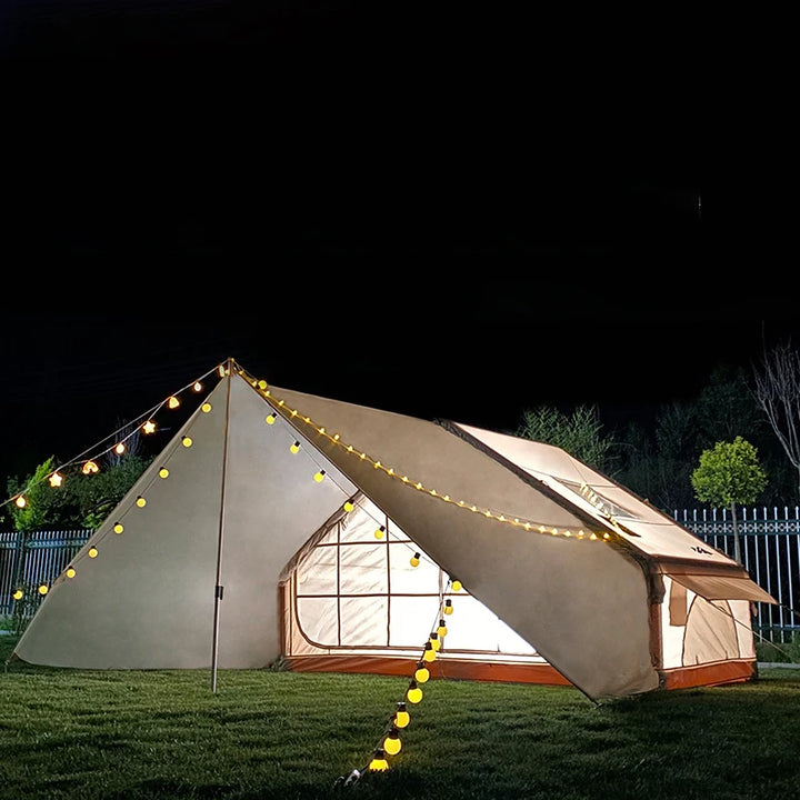 Une tente gonflable beige avec un grand auvent est installée dans l'herbe. Il fait nuit et il y a une guirlande lumineuse accrochée dessus. 