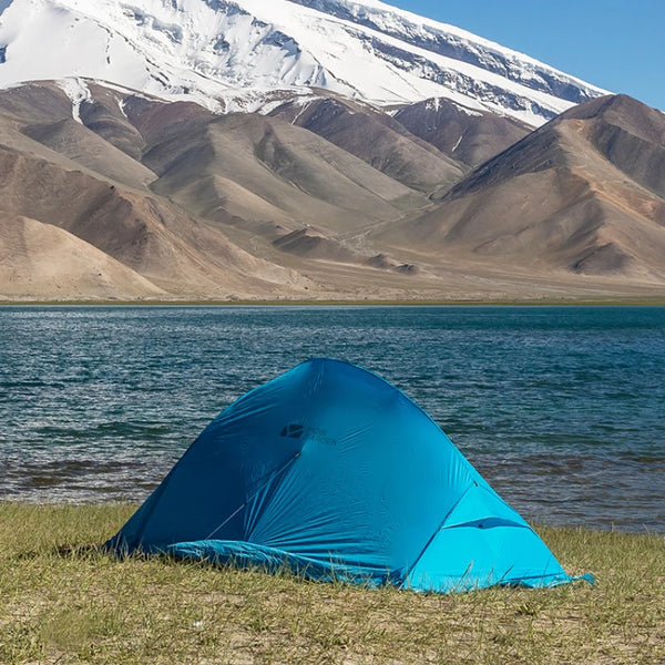 Une tente légère pour bivouac est installée au bord d'un lac de montagne. La tente est bleue. La montagne derrière est enneigée.