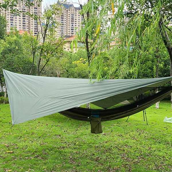 Une toile de tente verte est posée au dessus d'un hamac. La toile et le hamac sont installées dans un jardin. 