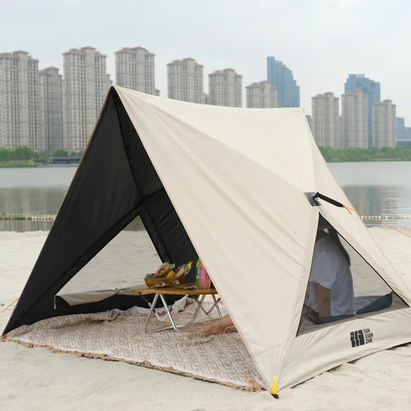 Une tente de plage canadienne beige est installée sur du sable. Elle a deux grandes ouvertes et des petites fenêtres sur chaque côté. 
