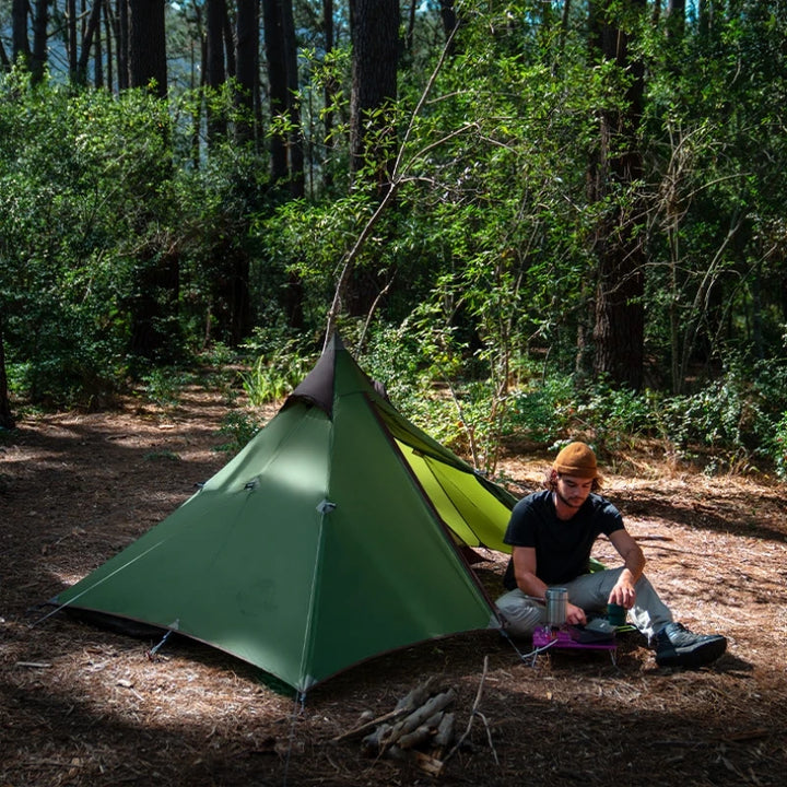 Un homme est dans la nature avec une tente tipi verte installée. Il est assis à côté. Il porte un bonnet marron, un tee-shirt noir et un pantalon gris.