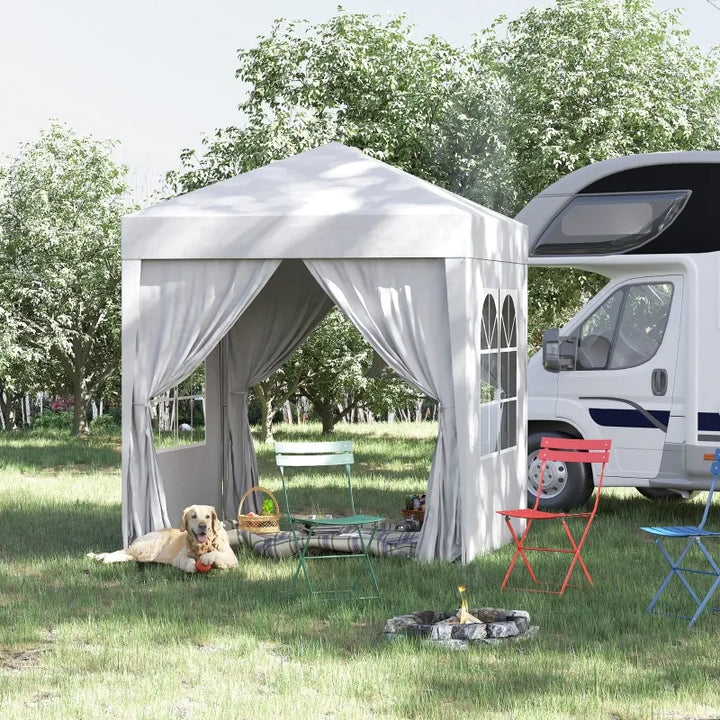 Une tente barnum blanche est installation dans l'herbe proche d'un camping car. La tente a une double ouverte avec deux pans à fenêtres sur les côtés. Il y a devant un chien et trois chaises. 