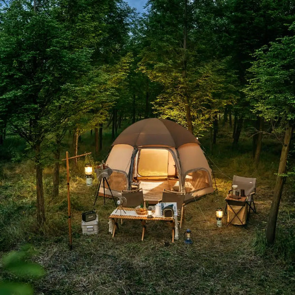 Une tente beige ronde est installée dans une forêt. Il y a devant une table, des chaises et de la décoration. 