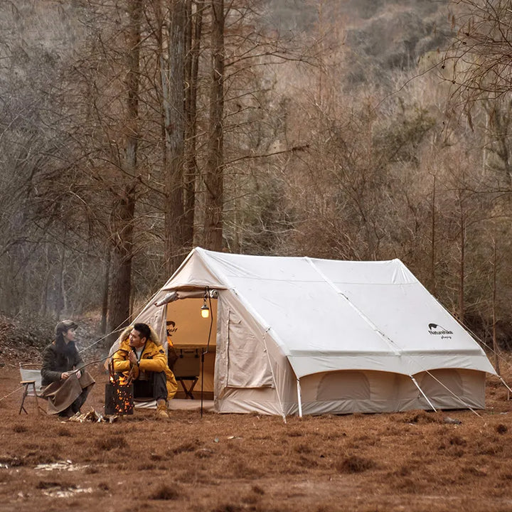 Une tente est posée dans la nature. C'est une tente gonflable beige pour 4 personnes. Il y a un homme et une femme assis devant. 