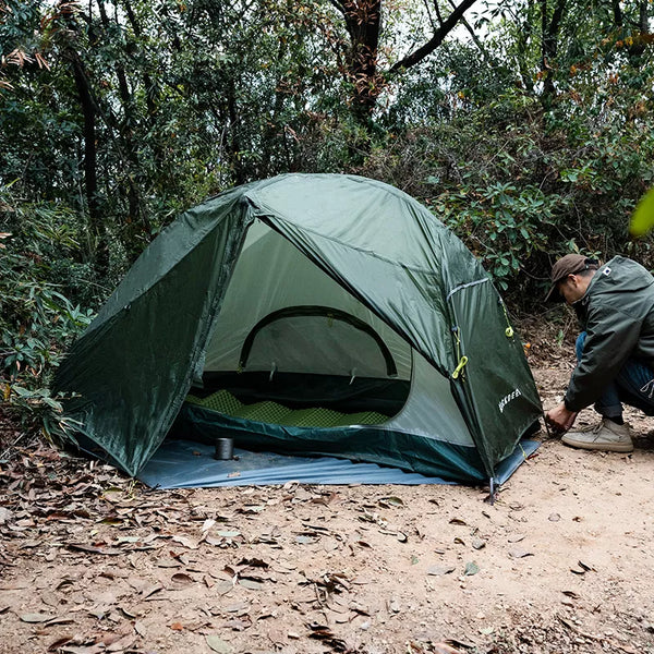 Une tente militaire pour bivouac ultra légère est en train d'être montée par un homme. La tente est verte militaire. Elle est dans la nature. 