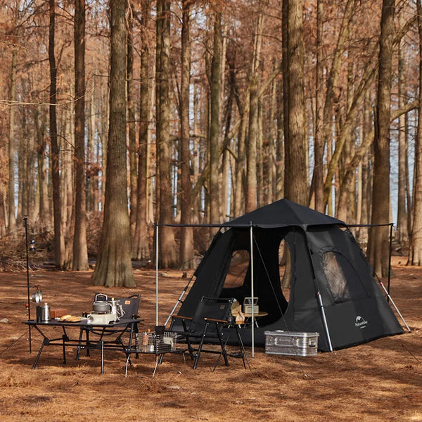 Une tente noire est dans une forêt. Il y a une table et des chaises devant. La tente a trois fenêtres et une grande porte. Il y a un abri devant la tente. 