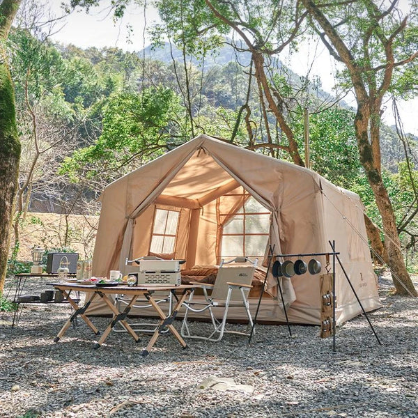 C'est une tente beige gonflable pour 3 personnes. C'est une tente lodge avec des fenêtres. Elle est dans la nature avec une table et des chaises devant. 