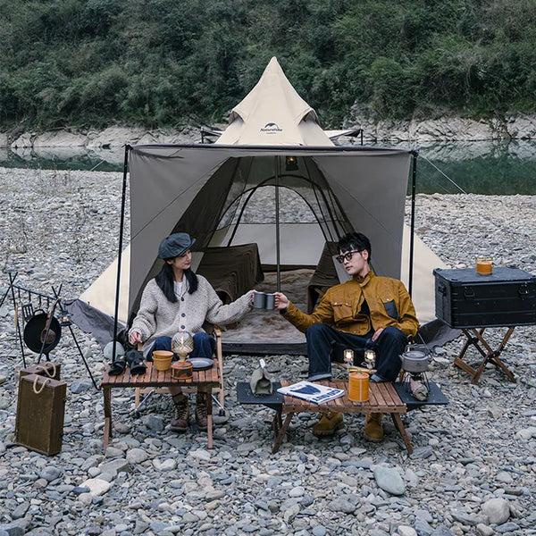 Une tente tipi beige est installée dans la nature au bord d'un étange. Il y a deux personnes, une table et deux fauteuils devant la tente.