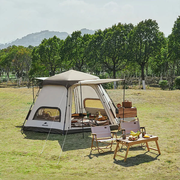 Une tente familiale et hexagonale est posée dans un parc. Elle est beige avec deux portes et de nombreuses fenêtres. Il y a devant la tente une table basse et deux fauteuils. 