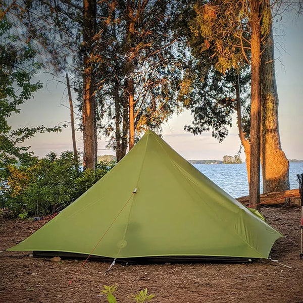 Une tente tipi vert pour bivouac est installé dans une forêt au bord de l'eau. 