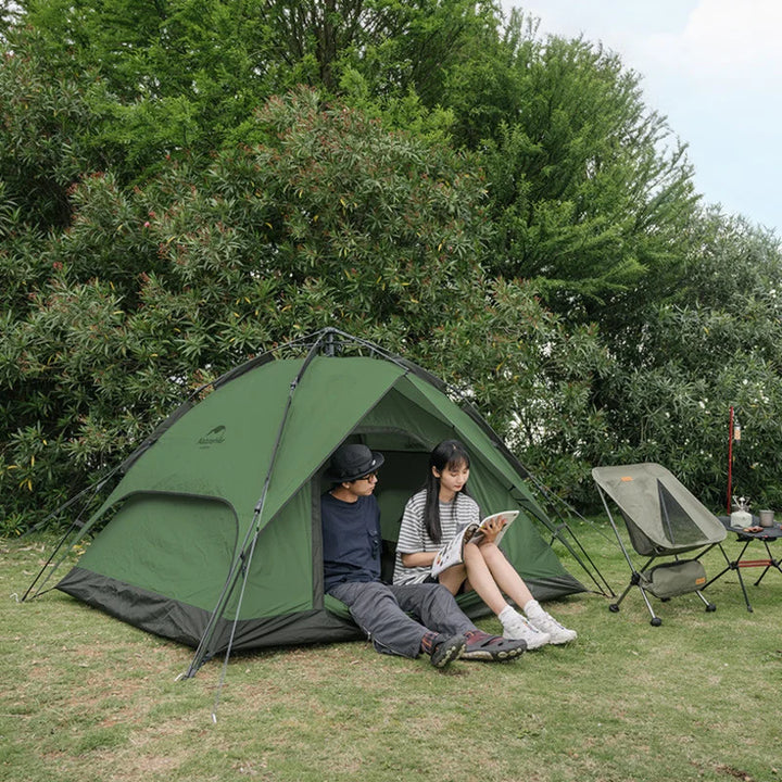 Une femme et un homme sont assis dans une tente couleur kaki. La tente est posée dans de l'herbe. Il y a un fauteuil et une petite table à côté. 