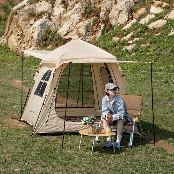 Une tente beige de glamping avec des fenêtres et une porte est installée dans la nature. Il y a une femme devant assise sur un fauteuil. Devant il y a une  une table basse. 