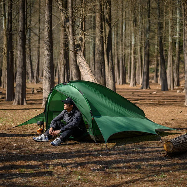 Une tente verte grande taille est installée dans la forêt. Un homme est assis devant. 