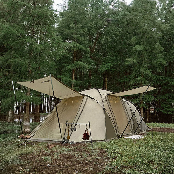 Une tente tunnel beige est installée dans la nature, en lisière de forêt. C'est une tente tunnel avec deux portes, une latérale et une sur l'extrémité. 