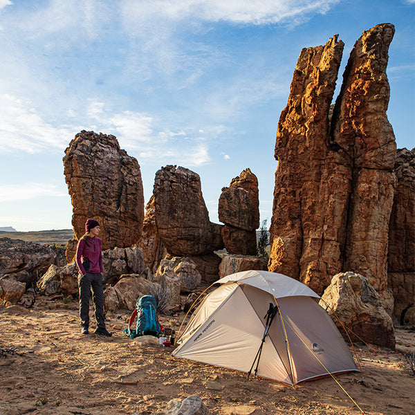 Une femme pose à côté d'une tente dans un environnement de désert américain. La tente est une tente de trekking légère beige et orange. 