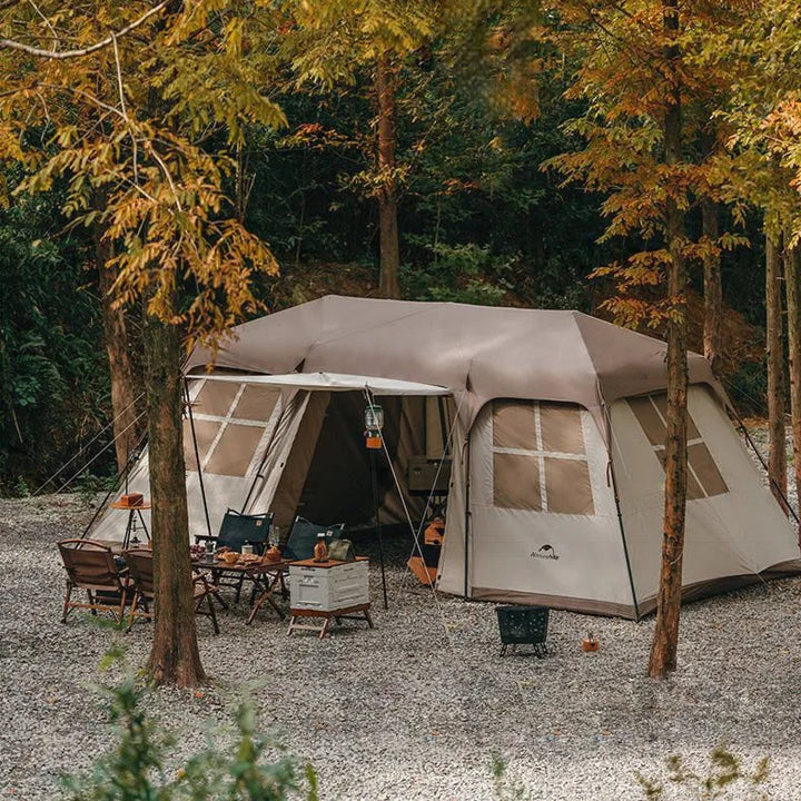 Une tente est installée dans la nature. Elle est beige et marron. Elle a un salon et de chaque côté une chambre. Il y a une table basse et des fauteuils devant la tente.