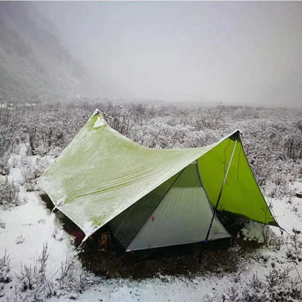 Une tente légère de trekking avec auvent vert est installée en pleine nature. Le sol est recouvert de neige. 