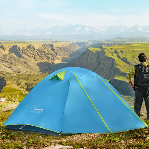Un homme est face à des montagnes à côté d'une tente. La tente est basique, pour une personne. Elle est bleu turquoise.