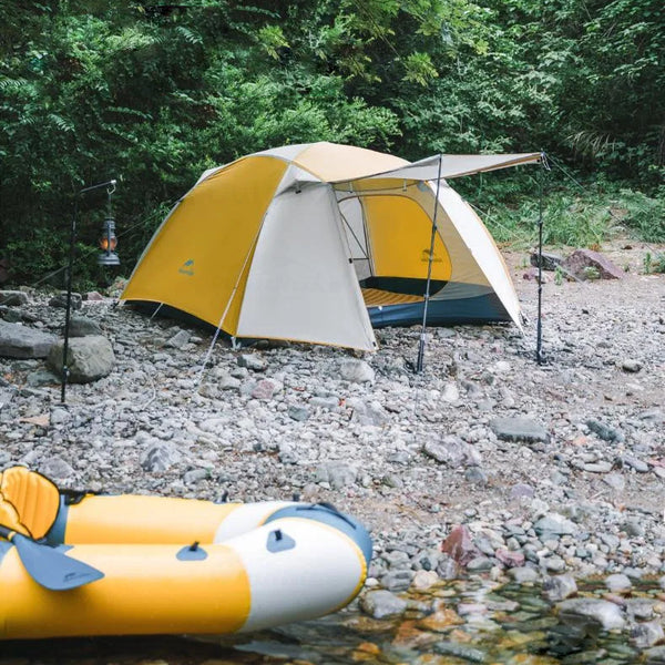Une tente blanche et jaune de trekking est posé au bord d'un ruisseau dans la nature sur des cailloux. 