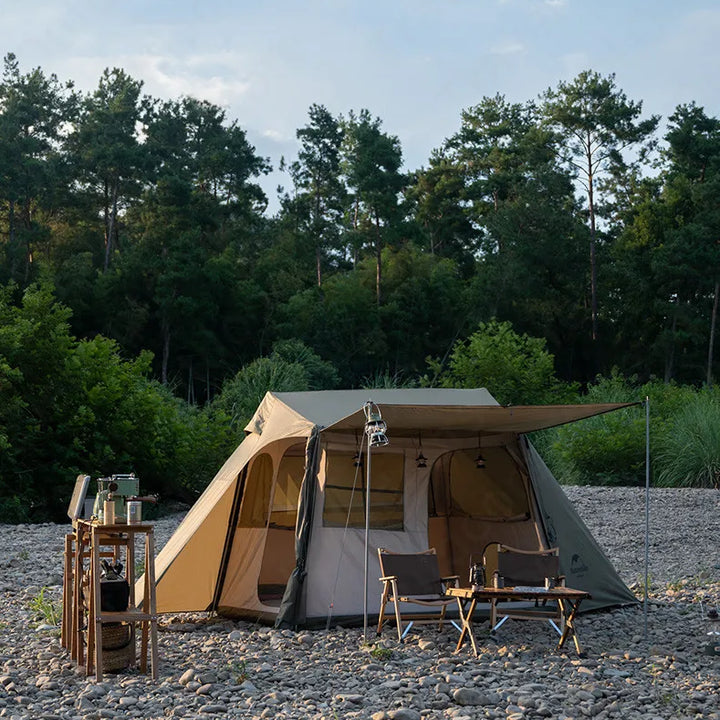 Une tente est posée sur des galets. Elle a une forme lodge et est kaki et beige. Elle a deux portes et de nombreuses fenêtres. Il y a devant la tente deux chaises et une table basse. À côté de la tente il y a une petite desserte qui fait office de cuisine. 